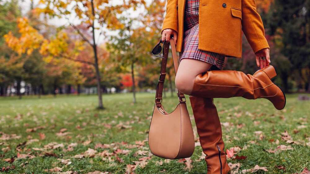 A woman poses in knee-high heeled cognac boots in front of a fall scene