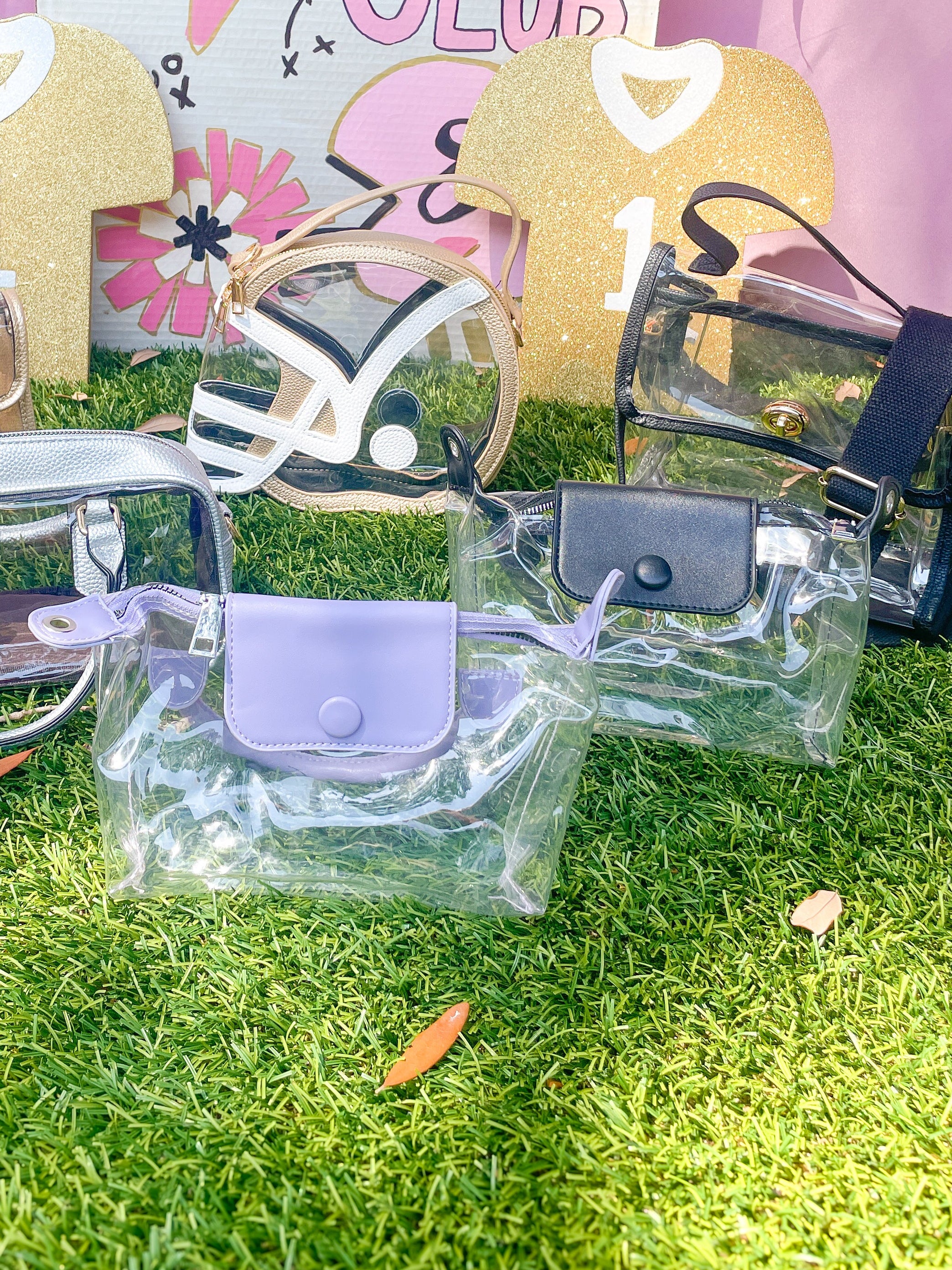 A collection of clear plastic handbags and small pouches displayed on artificial grass in front of a pink and white 'Swoon Sports Club' backdrop. The bags feature various colors including lavender, black, silver, and gold, with some showing their interior compartments through the transparent material.