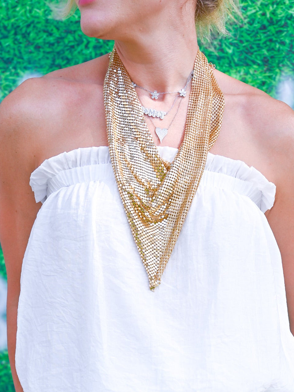 A woman wearing a white off-the-shoulder top and black-and-white zebra print pants, accessorized with a chunky gold chain necklace and bracelet, standing in front of a green textured backdrop with white numbers.