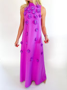 A sleeveless high-neck maxi dress in a vibrant magenta pink, decorated with 3D purple fabric flowers, shown on a model against a white studio background.