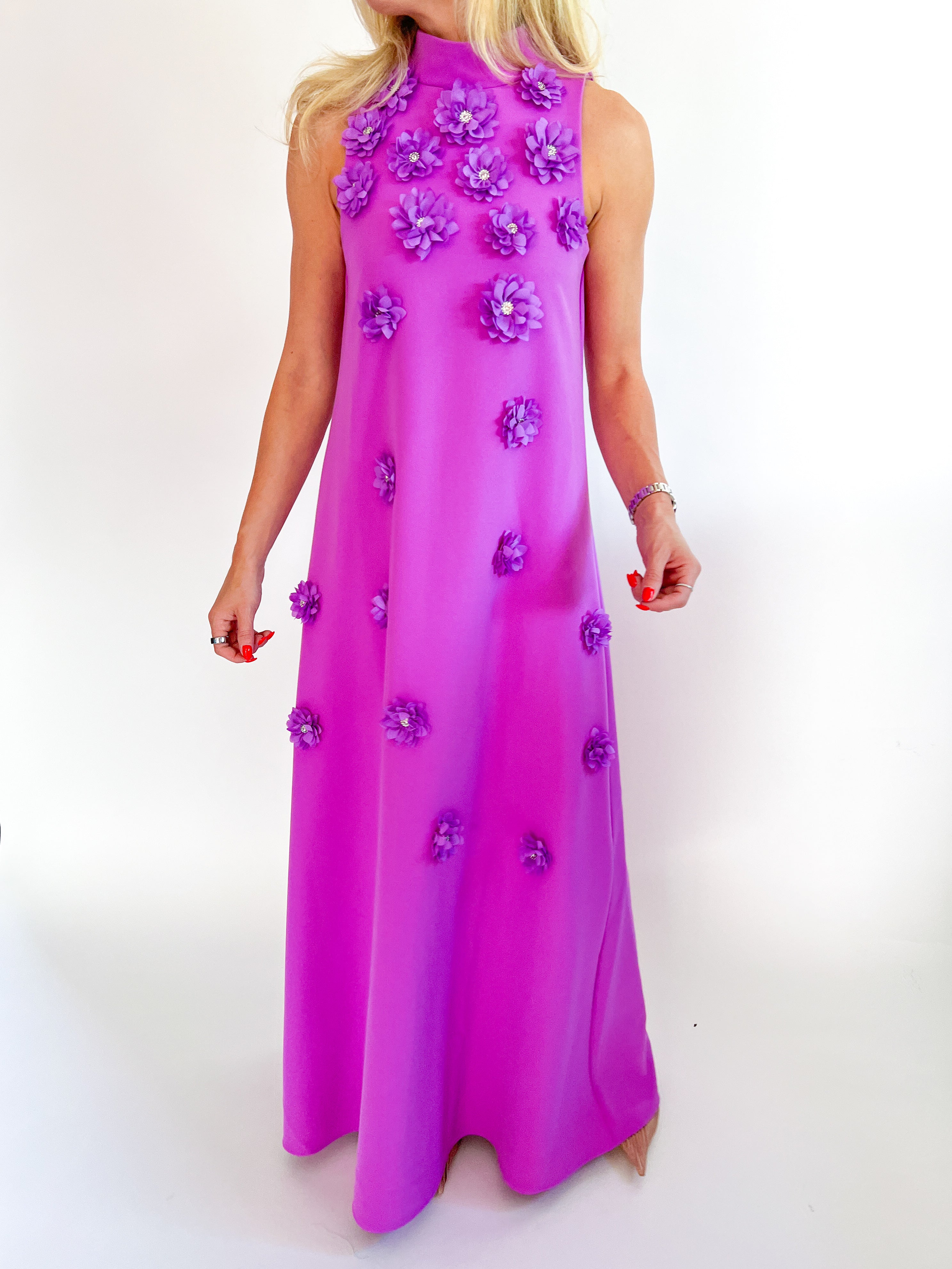 A sleeveless high-neck maxi dress in a vibrant magenta pink, decorated with 3D purple fabric flowers, shown on a model against a white studio background.