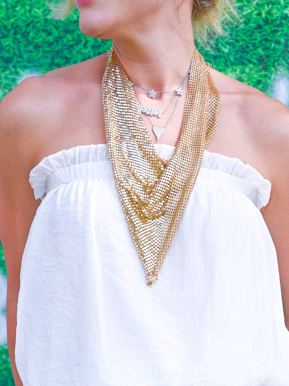 A woman wearing a white off-the-shoulder top and black-and-white zebra print pants, accessorized with a chunky gold chain necklace and bracelet, standing in front of a green textured backdrop with white numbers.