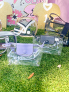 A collection of clear plastic handbags and small pouches displayed on artificial grass in front of a pink and white 'Swoon Sports Club' backdrop. The bags feature various colors including lavender, black, silver, and gold, with some showing their interior compartments through the transparent material.