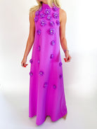 A sleeveless high-neck maxi dress in a vibrant magenta pink, decorated with 3D purple fabric flowers, shown on a model against a white studio background.