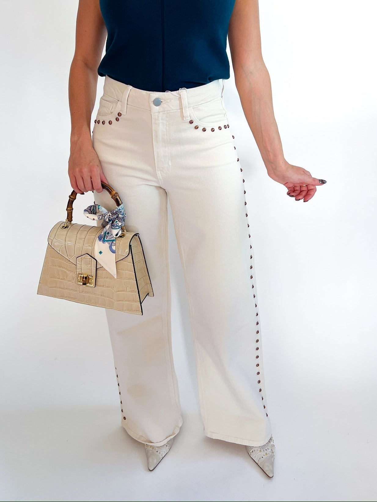 A model wearing a navy blue sleeveless top, off-white wide-leg jeans with silver studs, and holding a beige handbag, set against a plain white studio background.